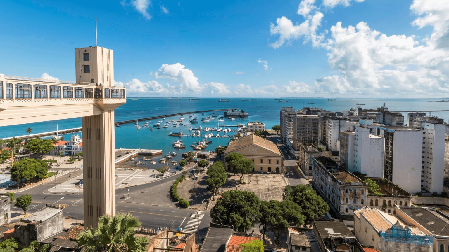Uma fotografia aérea panorâmica da Baía de Todos os Santos, em Salvador, Bahia, em um dia ensolarado. No primeiro plano, à esquerda, destaca-se a estrutura do Elevador Lacerda, com suas torres e cabines. A vista se estende pela cidade, mostrando prédios, ruas e árvores. No centro da imagem, a baía azul-turquesa, pontilhada por barcos e navios, se estende até o horizonte, onde se encontram o céu azul claro com nuvens brancas.