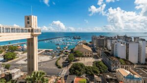 Uma fotografia aérea panorâmica da Baía de Todos os Santos, em Salvador, Bahia, em um dia ensolarado. No primeiro plano, à esquerda, destaca-se a estrutura do Elevador Lacerda, com suas torres e cabines. A vista se estende pela cidade, mostrando prédios, ruas e árvores. No centro da imagem, a baía azul-turquesa, pontilhada por barcos e navios, se estende até o horizonte, onde se encontram o céu azul claro com nuvens brancas.