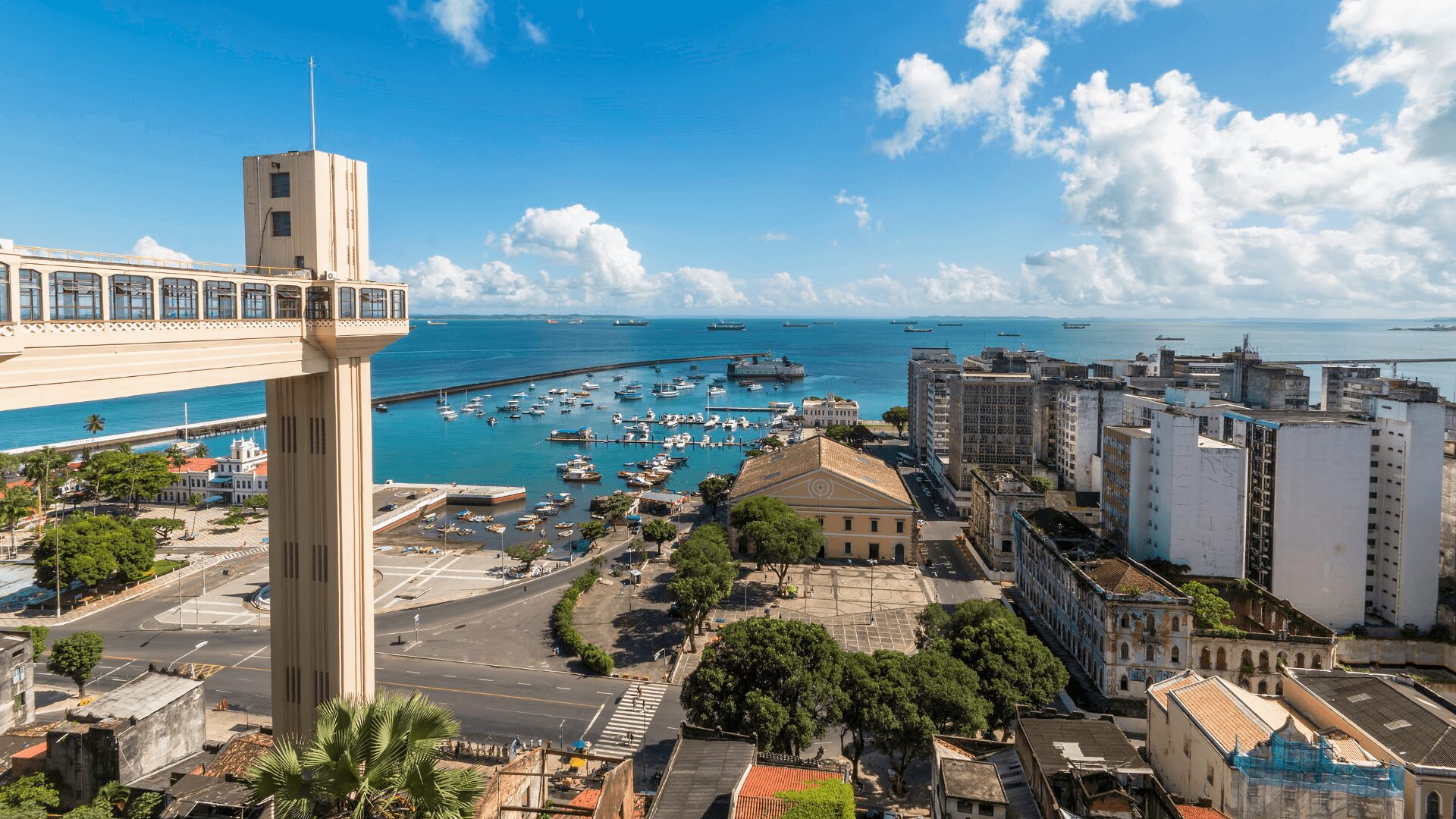 Uma fotografia aérea panorâmica da Baía de Todos os Santos, em Salvador, Bahia, em um dia ensolarado. No primeiro plano, à esquerda, destaca-se a estrutura do Elevador Lacerda, com suas torres e cabines. A vista se estende pela cidade, mostrando prédios, ruas e árvores. No centro da imagem, a baía azul-turquesa, pontilhada por barcos e navios, se estende até o horizonte, onde se encontram o céu azul claro com nuvens brancas.