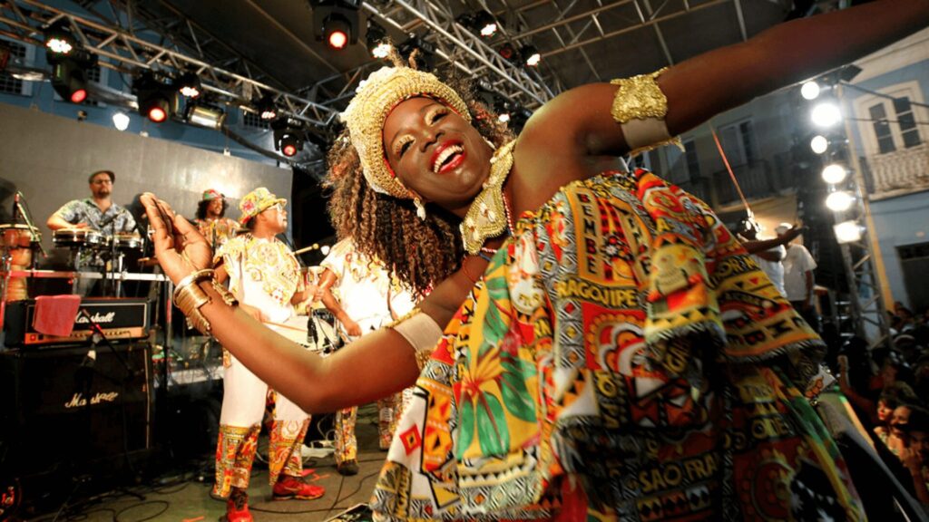 Um artista, com trajes tradicionais vibrantes, dança energicamente no palco, cercado por músicos e luzes coloridas.