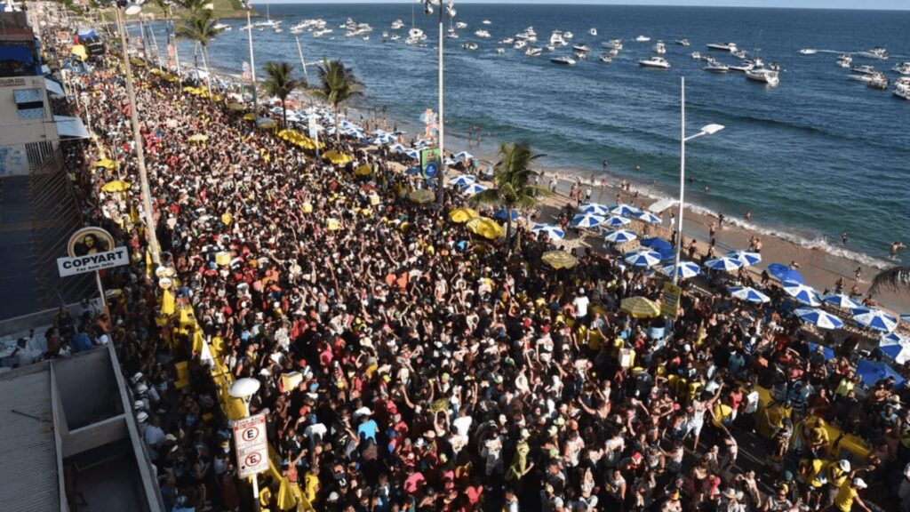 Uma multidão vibrante se reúne ao longo de um calçadão à beira-mar, com guarda-sóis e barcos visíveis no oceano, criando um cenário costeiro animado.