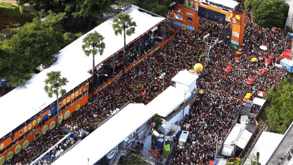 Vista aérea de uma grande multidão reunida em um evento festivo, cercada por barracas coloridas e palmeiras.