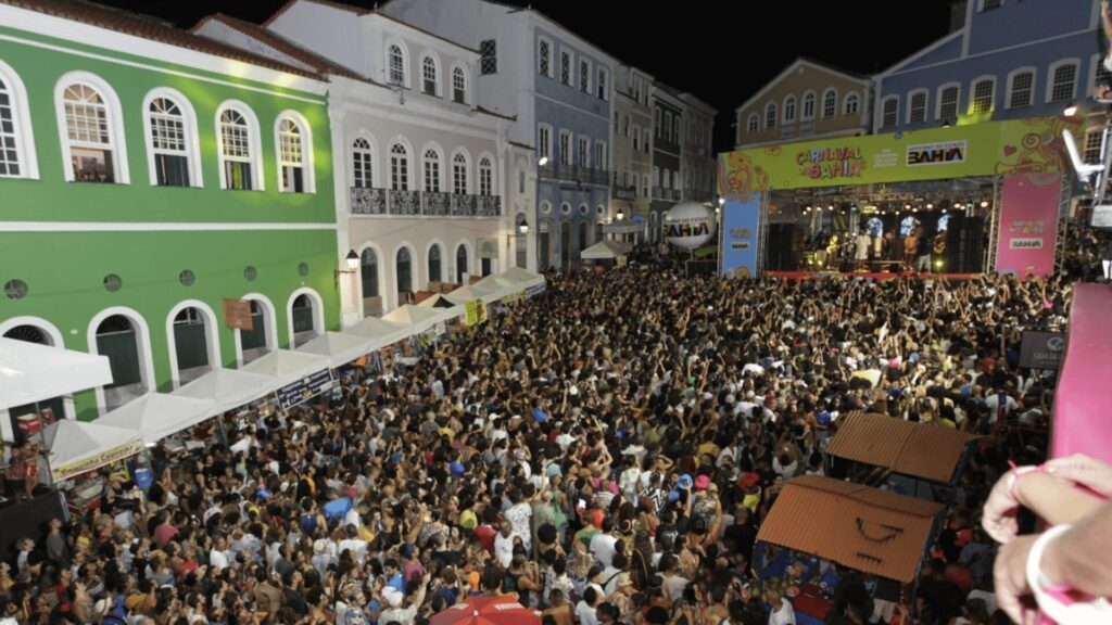 Uma multidão vibrante curtindo um festival noturno, prédios coloridos ao fundo, palco e vendedores visíveis em Salvador, Brasil.