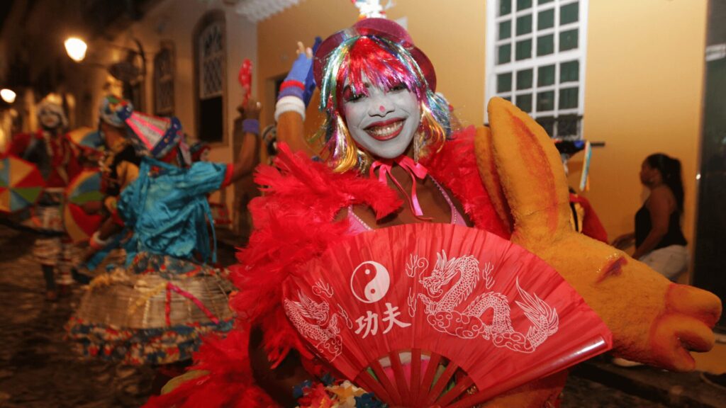 Um artista vibrante em trajes coloridos segura um leque e fica ao lado de outras pessoas em um animado carnaval de rua.