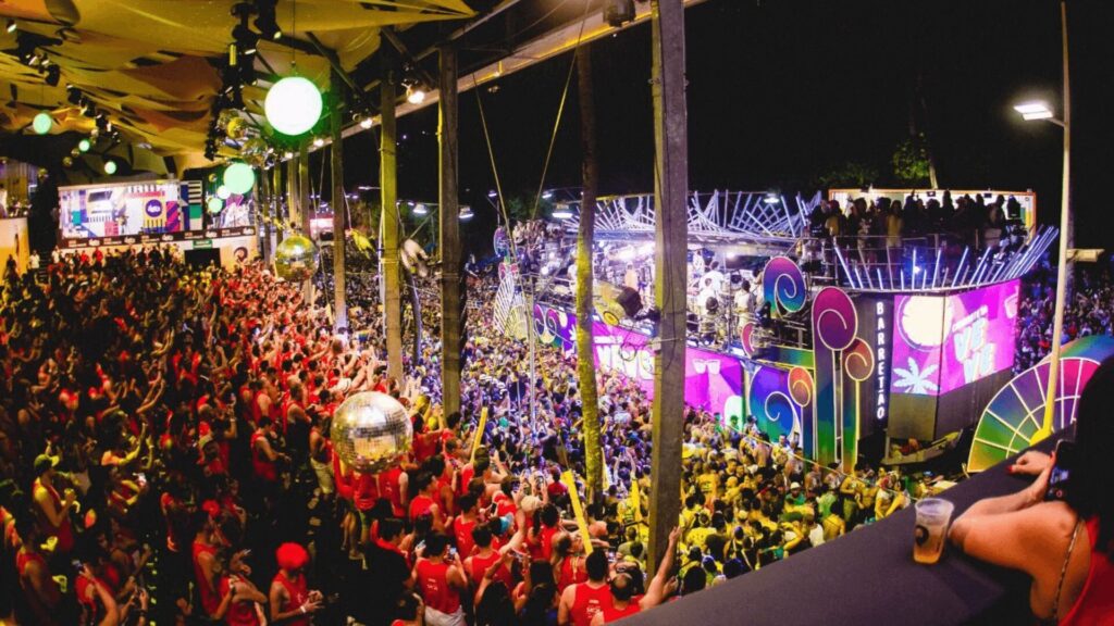 Um cenário vibrante de festival noturno com uma enorme multidão em camisas vermelhas, luzes coloridas, bolas de discoteca e um palco animado ao fundo.