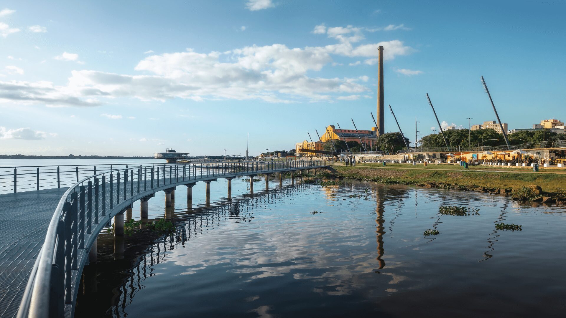 Uma passarela curva ao longo de um rio refletindo o céu azul, com um edifício industrial, mastros e vendedores alinhados à margem.