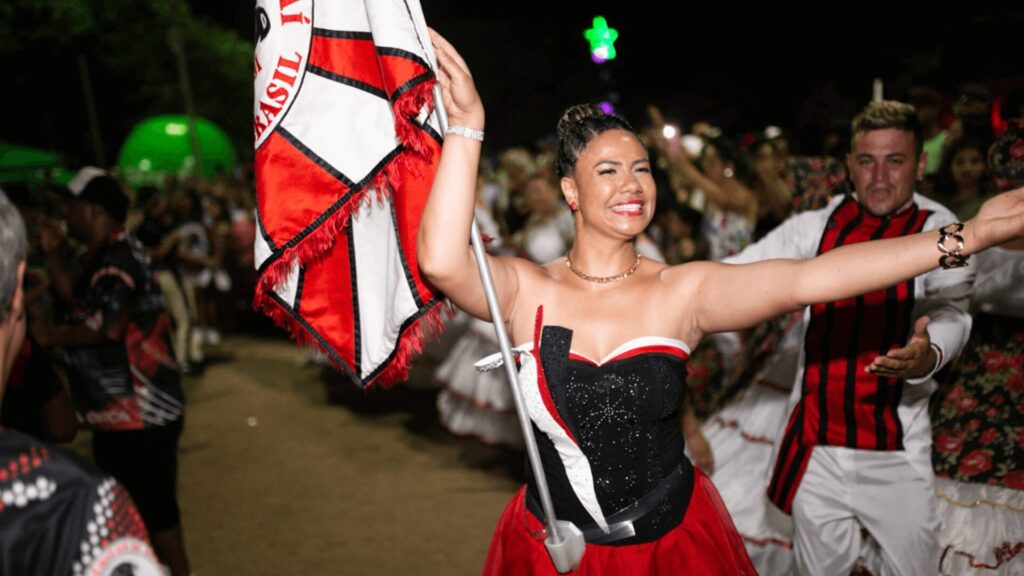 Uma mulher com um vestido festivo agita apaixonadamente uma bandeira durante uma animada celebração, cercada por uma multidão em trajes coloridos.