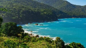 Montanhas verdejantes flanqueiam um mar azul-turquesa tranquilo, com ondas suaves batendo nas costas rochosas sob um céu azul claro.