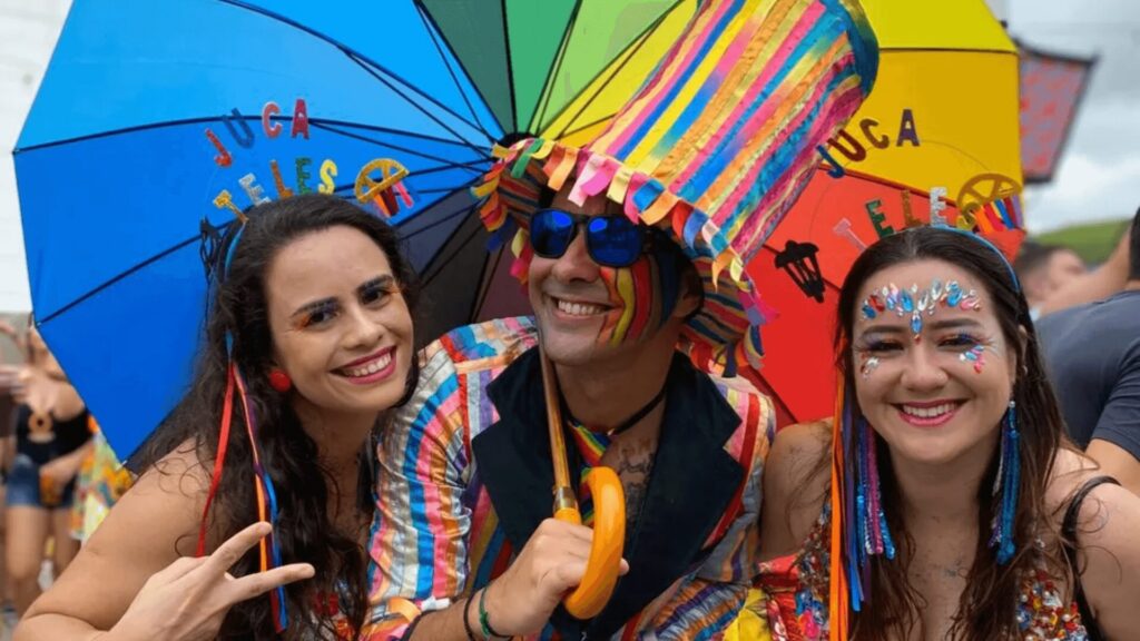 Três amigos em trajes vibrantes posam alegremente com guarda-chuvas coloridos em um evento festivo ao ar livre.