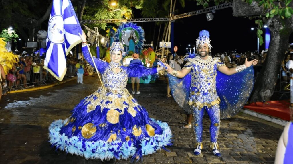 Uma vibrante cena de carnaval com dançarinos em deslumbrantes trajes azuis e dourados, comemorando com uma bandeira sob luzes festivas.