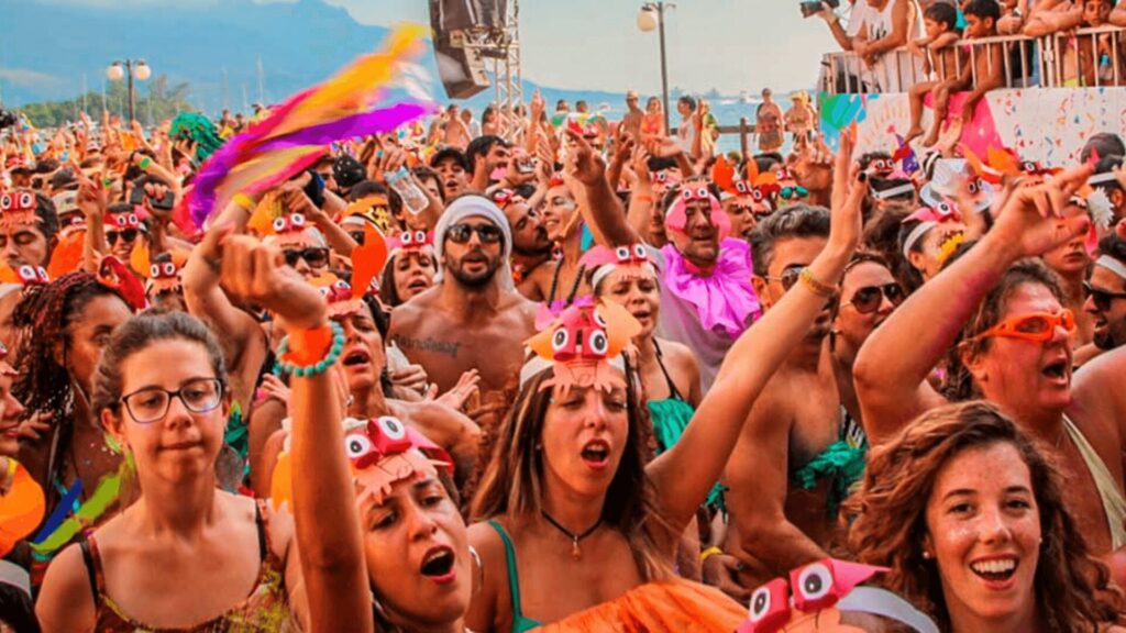 Uma multidão vibrante em um festival, usando chapéus e acessórios coloridos, dançando alegremente com as mãos levantadas em um cenário cênico.