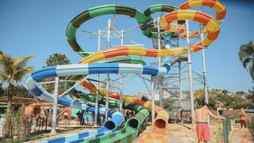 Um parque aquático colorido com escorregadores sinuosos, uma escadaria alta e visitantes aproveitando o dia ensolarado sob um céu azul claro.