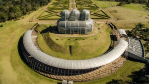 Fotografia aérea do Jardim Botânico de Curitiba, com destaque para a icônica estufa de três cúpulas de vidro, o grande semicírculo de estrutura metálica, os jardins simétricos dispostos em padrões geométricos e o lago com chafariz.