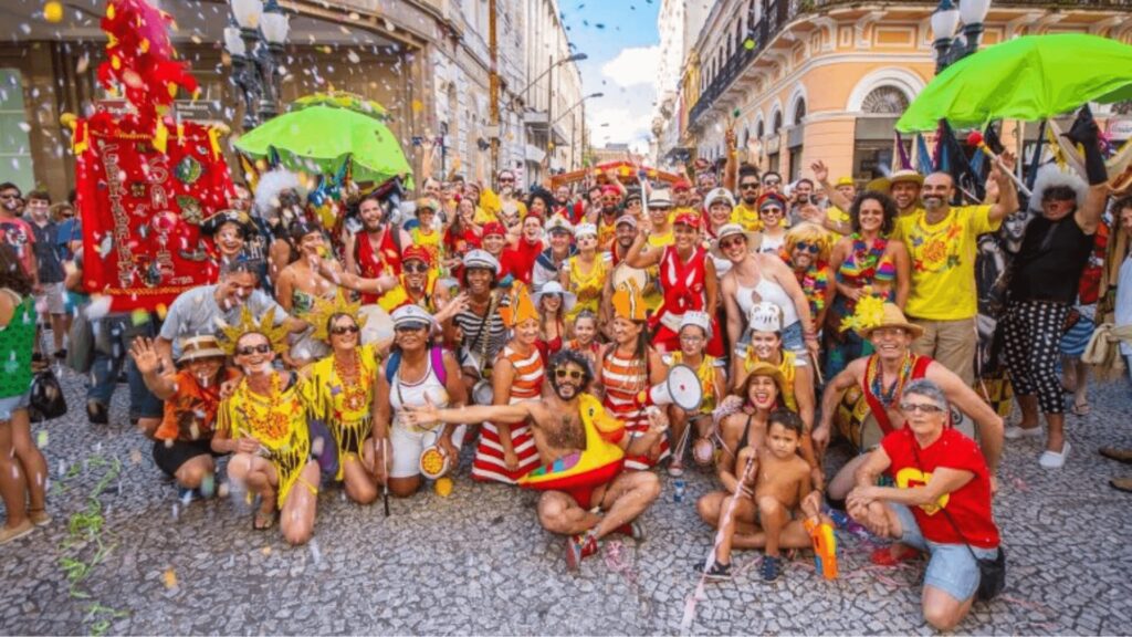 Uma festa animada de um grupo durante um festival, com fantasias coloridas, confetes e gente feliz posando em uma rua movimentada.