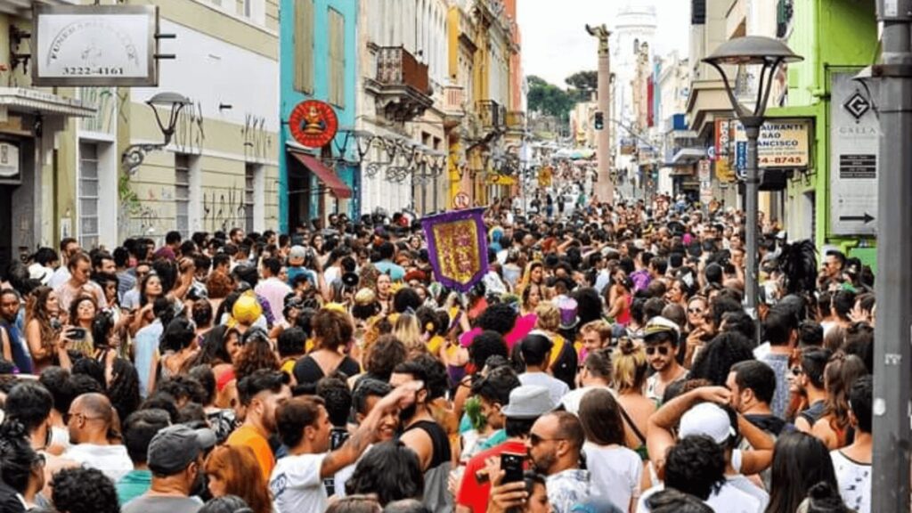 Uma multidão animada toma conta de uma rua colorida durante uma festa, com pessoas vestindo roupas coloridas e segurando enfeites.