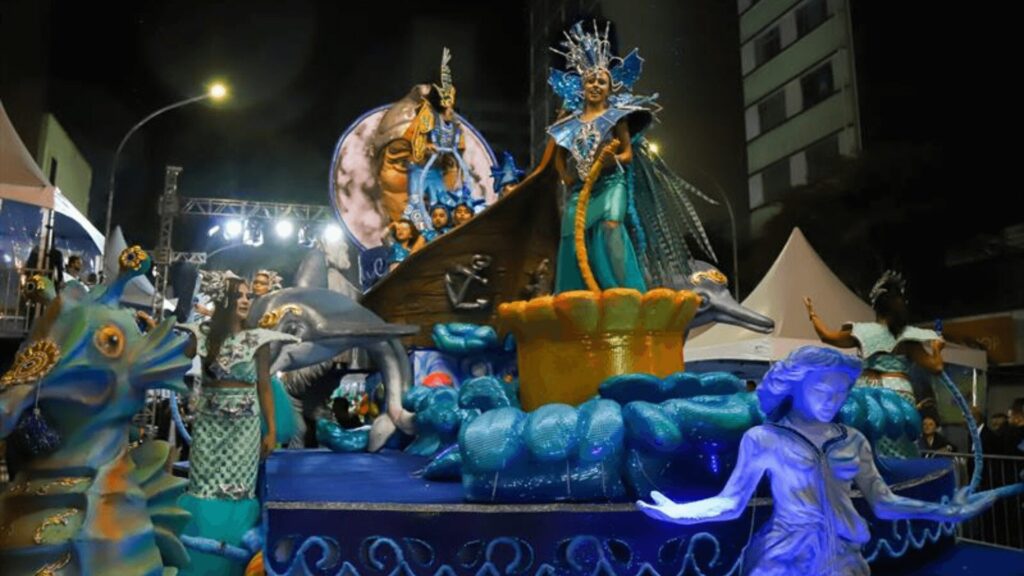 Um carro alegórico de carnaval super colorido, enfeitado com bichos do mar e gente fantasiada, celebrando a vida marinha debaixo das luzes da festa, à noite.