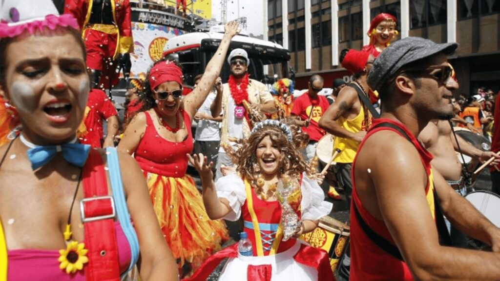 Uma festa de rua animada, com fantasias coloridas, dançarinos, batuqueiros e confetes, mostrando toda a alegria da celebração.