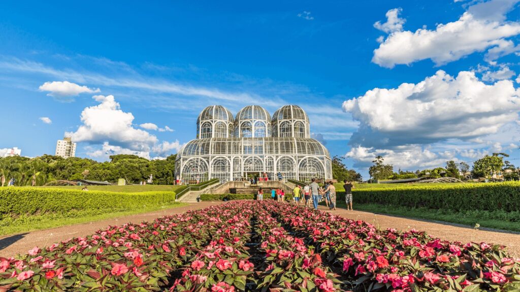 Uma estufa de vidro com teto arredondado fica em um parque lindo, cheio de flores rosas e gente aproveitando o sol.