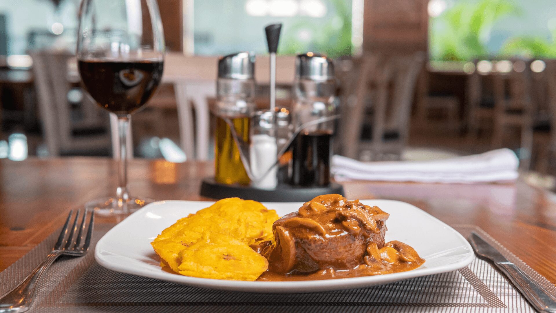 Um prato de carne tenra ao molho de cogumelos acompanhado de banana dourada frita, com uma taça de vinho tinto em ambiente de restaurante.