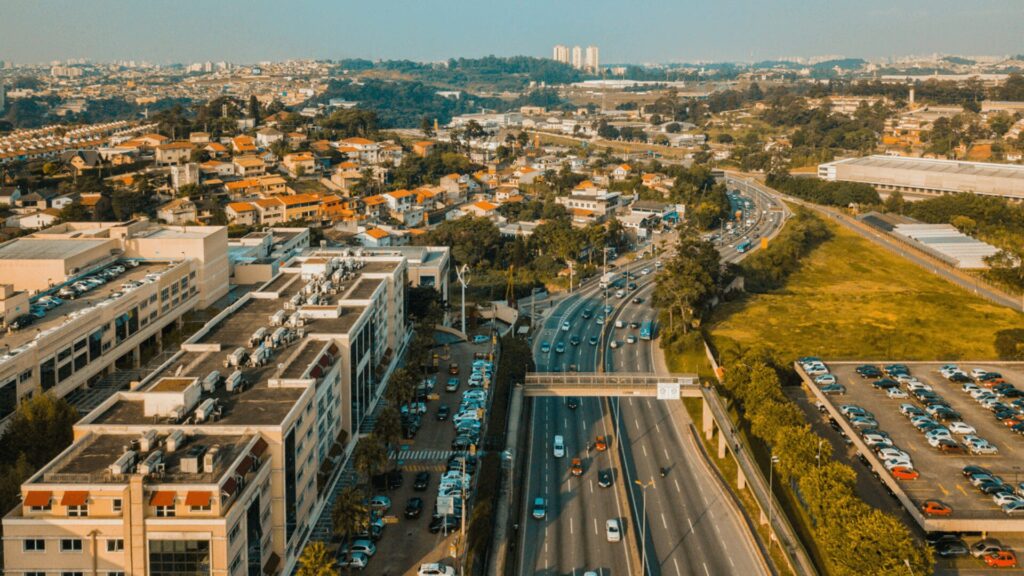 Uma vista aérea de uma rodovia movimentada cercada por áreas urbanas e suburbanas, apresentando trânsito e arquitetura diversificada.