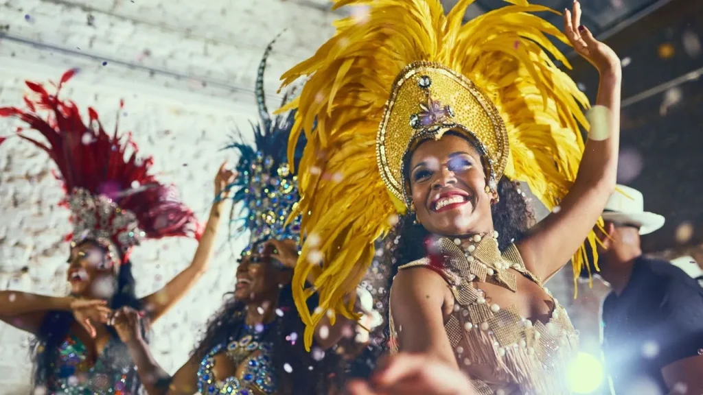 Close-up de uma passista de Carnaval sorrindo com os braços levantados em meio a confetes. Ela usa um imponente adereço de cabeça com penas amarelas volumosas e detalhes dourados. Ao fundo, outras passistas com fantasias em tons de vermelho e azul aparecem desfocadas, reforçando o clima de festa. A iluminação é quente e destaca o brilho das fantasias.