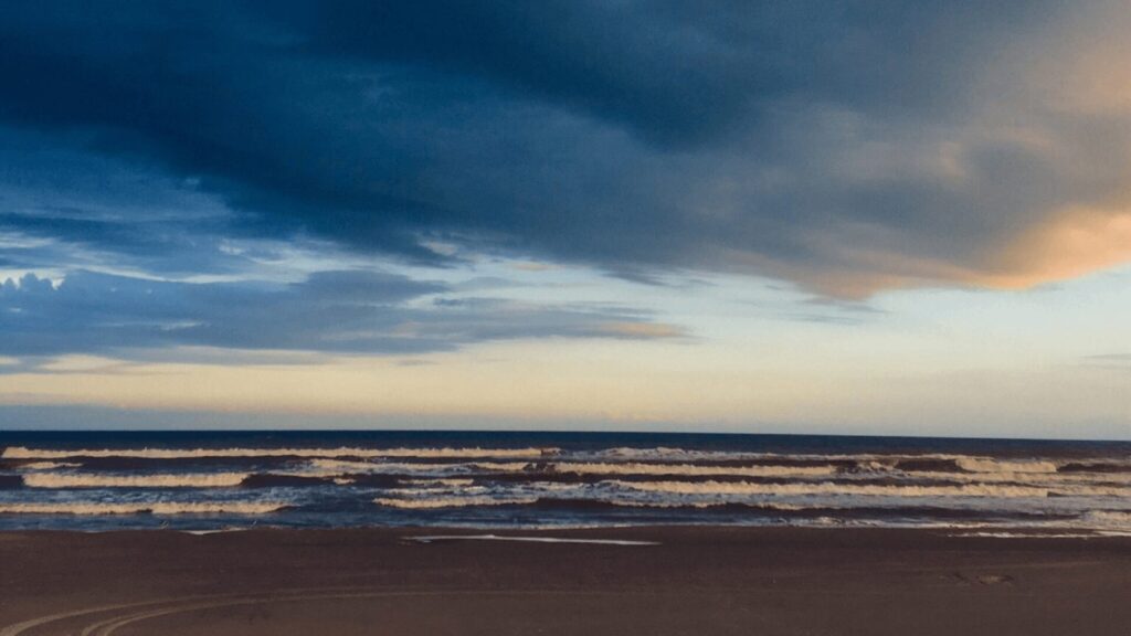 Uma cena serena de praia ao anoitecer, com ondas ondulantes sob um céu dramático com nuvens escuras e toques de cores pastel do pôr do sol.
