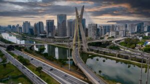 Vista aérea da icônica ponte e do horizonte de São Paulo, refletidos no rio sob um céu dramático. Estradas movimentadas com tráfego abaixo.