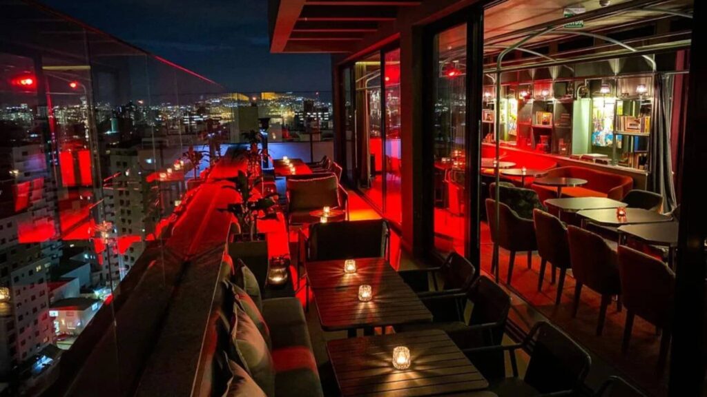 A imagem retrata o ambiente de um bar sofisticado localizado no terraço (rooftop) de um edifício, durante a noite. A iluminação predominante é vermelha e baixa, criando uma atmosfera íntima e moderna.
No primeiro plano e à esquerda, há uma área externa com um guarda-corpo de vidro. Mesas e sofás com almofadas estão dispostos neste espaço, com pequenas velas acesas sobre as mesas, adicionando um toque acolhedor. Ao fundo desta área, é possível ver as luzes de uma cidade à noite, com edifícios iluminados e, mais distantemente, uma estrutura que se assemelha a um monumento antigo iluminado em uma colina. O céu está escuro.
À direita e no interior do bar, separado por grandes janelas de vidro, o ambiente continua com a mesma iluminação avermelhada. Vê-se mesas redondas e cadeiras, além de um balcão ou área de bar ao fundo, com prateleiras iluminadas exibindo garrafas. A decoração interna parece incluir elementos artísticos ou quadros nas paredes.
A perspectiva da foto é de dentro para fora, capturando tanto o ambiente interno quanto a vista panorâmica da cidade a partir do terraço. A sensação geral é de um local elegante e com vista privilegiada.