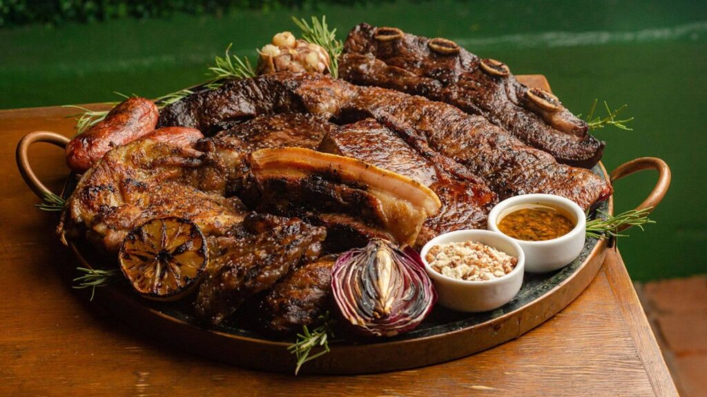A imagem exibe, em primeiro plano, uma grande travessa redonda de metal, possivelmente cobre ou ferro, com alças laterais, repleta de uma variedade de carnes assadas, estilo parrilla. A travessa está sobre uma superfície de madeira.
As carnes apresentam diferentes cortes e texturas, todas com aspecto suculento e bem grelhado. É possível identificar peças maiores como costela bovina com osso, cortes de bife mais grossos, e o que parece ser barriga de porco com a pele pururucada ao centro. Há também linguiças toscanas assadas, algumas com a pele mais tostada.
Espalhados entre as carnes, há elementos que adicionam aroma e sabor: uma cabeça de alho inteira assada, ramos de alecrim fresco, uma cebola roxa cortada ao meio e grelhada, e uma metade de limão também grelhado com a face cortada para baixo.
À direita da travessa, dois pequenos potes brancos contêm acompanhamentos: um com um molho amarelado e oleoso, possivelmente vinagrete ou chimichurri, e outro com farofa ou um tempero granulado.
Ao fundo, de forma desfocada, percebe-se uma superfície verde escura, que pode ser uma parede ou vegetação, e um brilho que sugere a presença de água, como uma piscina ou um lago. A iluminação da cena realça as cores e texturas das carnes, tornando a apresentação bastante apetitosa. A foto é tirada de um ângulo ligeiramente superior, oferecendo uma visão clara de todo o conteúdo da parrilla.