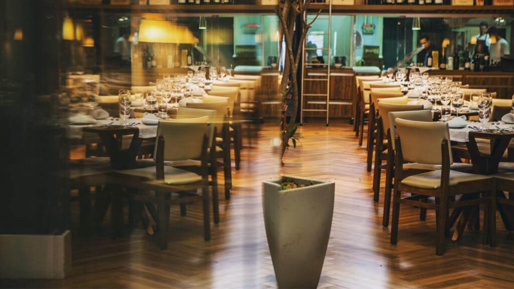 A imagem mostra o interior de um restaurante com um ambiente sofisticado e acolhedor. O ponto de vista é a partir da entrada ou de uma seção do salão, olhando para dentro.
O piso é de madeira clara, com um padrão em zigue-zague (espinha de peixe). O salão é preenchido por diversas mesas quadradas e retangulares, todas postas com toalhas brancas, taças de vinho, copos e talheres, prontas para receber os clientes. As cadeiras são de madeira com assentos e encostos estofados em um tom claro, bege ou creme.
No centro da imagem, ligeiramente em primeiro plano, há um vaso de plantas alto e moderno, de formato geométrico e cor clara. Mais ao fundo, no centro do salão, destaca-se o tronco de uma árvore ou um arranjo de galhos secos que se estende em direção ao teto, adicionando um elemento natural e decorativo ao espaço.
Ao fundo, é possível ver uma longa prateleira ou estante de madeira escura que ocupa grande parte da parede, repleta de garrafas de vinho e outras bebidas, sugerindo uma adega ou um bar bem abastecido. A iluminação é suave e indireta, com pendentes e focos de luz que criam uma atmosfera quente e convidativa. Reflexos de luz são visíveis nas superfícies polidas e nos vidros.
Em alguns pontos, há divisórias de vidro ou espelhos que ampliam o ambiente e refletem as luzes e as mesas. No canto superior direito, é possível entrever uma pessoa de uniforme branco, possivelmente um funcionário do restaurante. A profundidade de campo é relativamente curta, com o primeiro plano (o vaso) mais nítido e o fundo levemente desfocado, o que confere um ar artístico à fotografia.