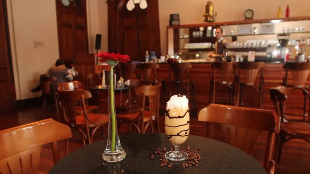 Uma mesa de café estilosa com um copo alto de café gelado coberto com chantilly, ao lado de um vaso com uma flor vermelha.