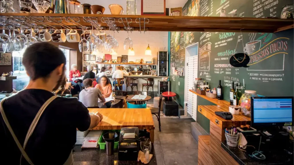 Um aconchegante café com mesas de madeira, copos pendurados e clientes apreciando bebidas e refeições em uma atmosfera vibrante.