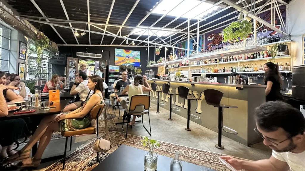 Um café animado com prateleiras de garrafas coloridas, clientes jantando em mesas e um mural vibrante adornando a parede.