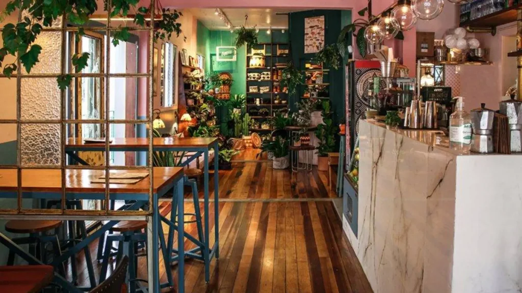 Um café aconchegante com piso de madeira, plantas verdes e decoração colorida, com um balcão vibrante e prateleiras cheias de produtos artesanais.