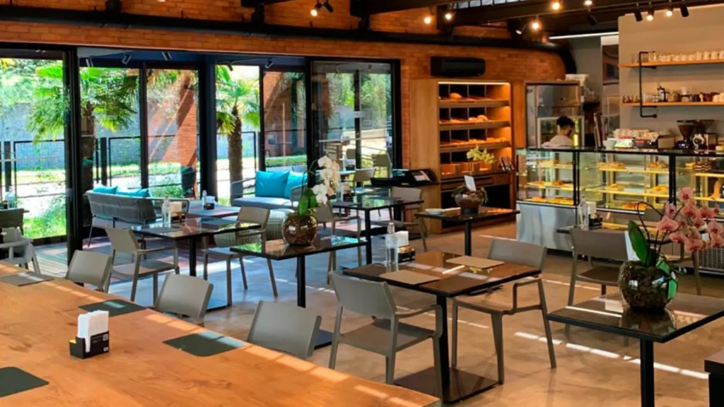 Interior moderno de um café com mesas de madeira, vitrines de vidro e muito verde visível através de grandes janelas.