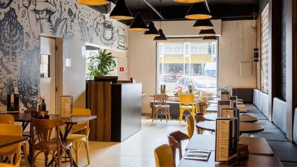 Um interior de café elegante com mesas de madeira, cadeiras amarelas, arte de parede excêntrica e grandes janelas que deixam entrar luz natural.