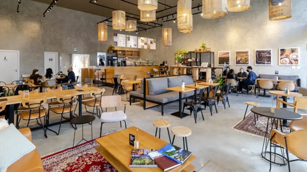 Um interior de café moderno com mesas e cadeiras de madeira, iluminação pendente e plantas, repleto de clientes aproveitando o tempo.