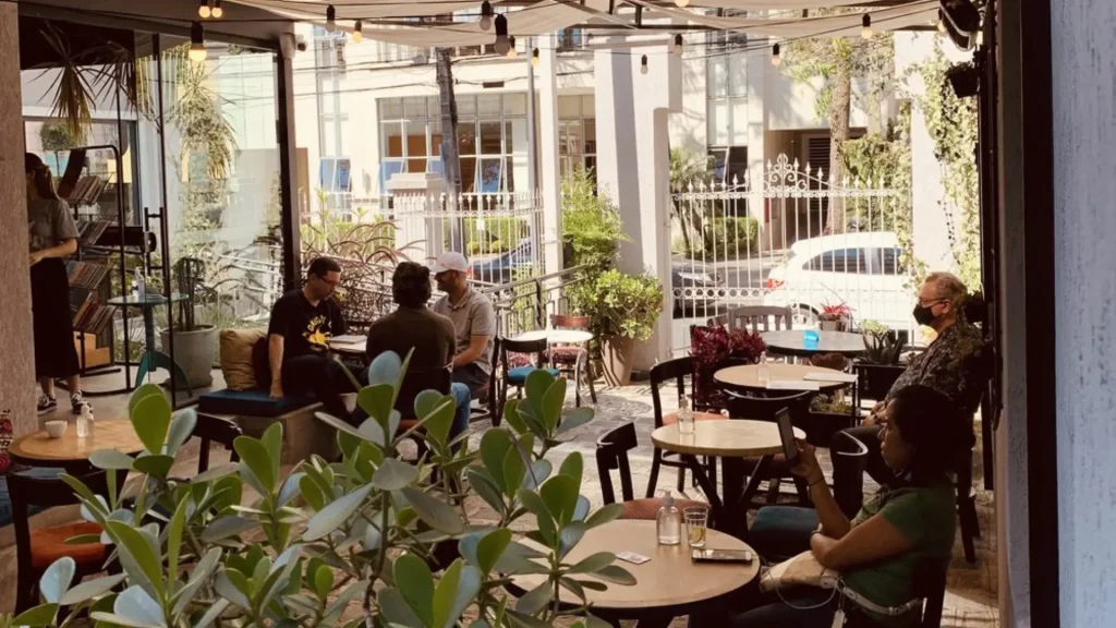 Um animado café com clientes sentados ao redor de mesas de madeira, cercados por vegetação e luz do sol filtrada por uma cobertura.
