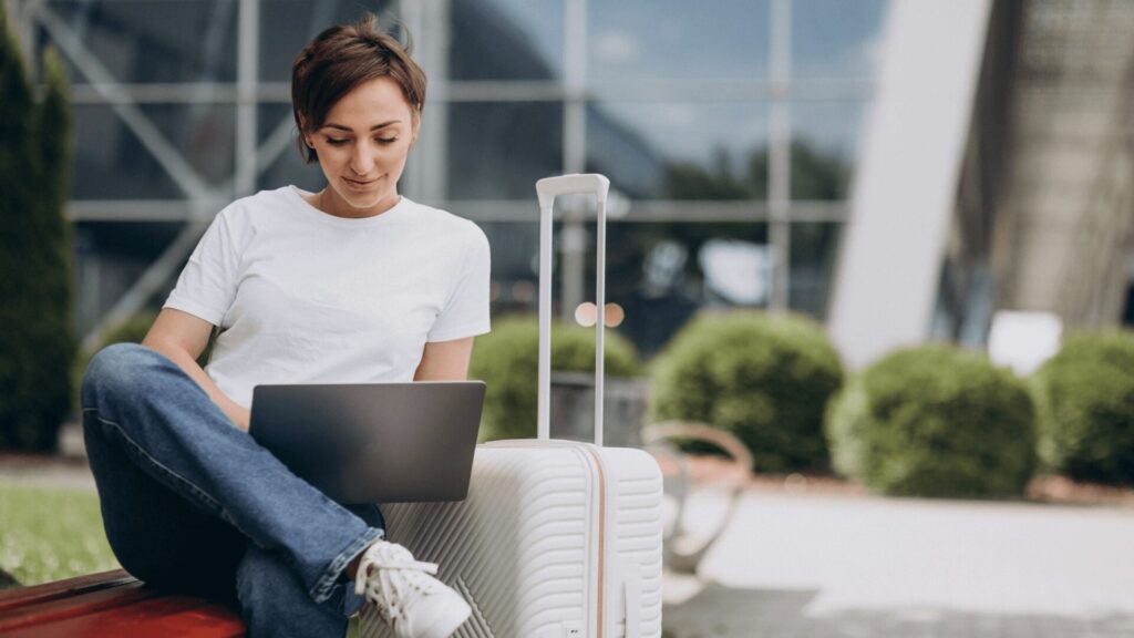 Uma pessoa sentada em uma mala vermelha usa um laptop em um ambiente externo vibrante com arbustos verdes e um edifício moderno ao fundo.