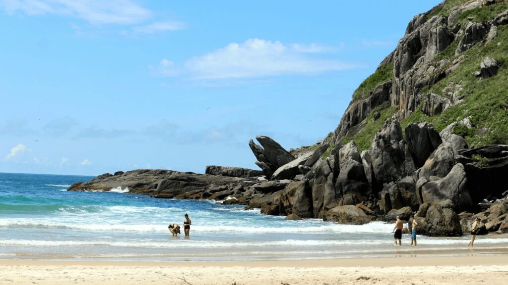 Duas mulheres brincando perto da água e dois homens em pé nas ondas, com penhascos rochosos e um céu azul claro ao fundo.