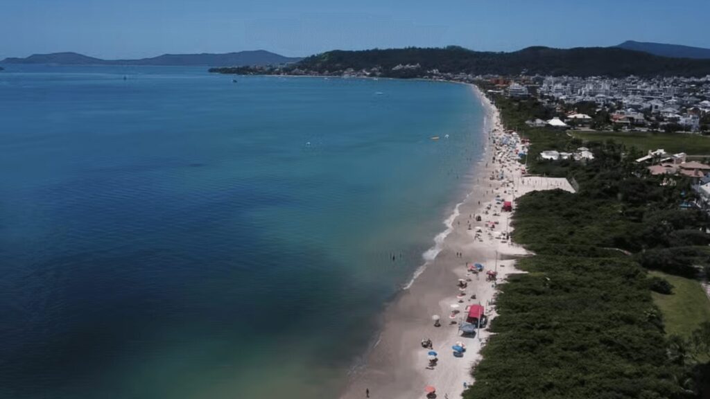 Vista aérea de uma praia de areia com guarda-sóis coloridos, água azul cristalina e montanhas distantes, movimentada com banhistas aproveitando o sol.