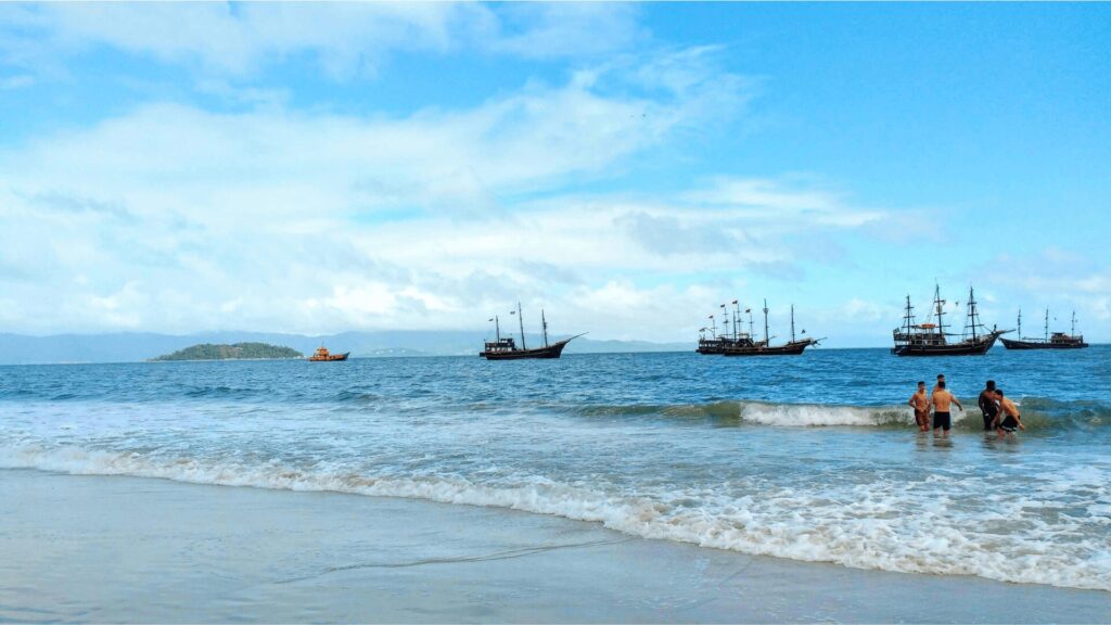 Duas pessoas caminham no oceano perto de uma praia arenosa, enquanto navios tradicionais navegam ao fundo sob um céu azul parcialmente nublado.