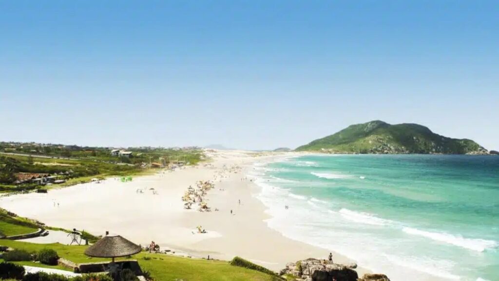 Uma praia pitoresca com areia dourada e ondas azul-turquesa, repleta de banhistas e guarda-sóis, tendo como pano de fundo colinas verdejantes.