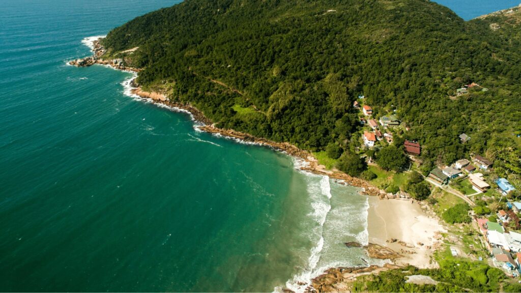 Vista aérea de uma praia tranquila cercada por vegetação exuberante e penhascos rochosos, com pequenas casas aninhadas ao longo da costa.