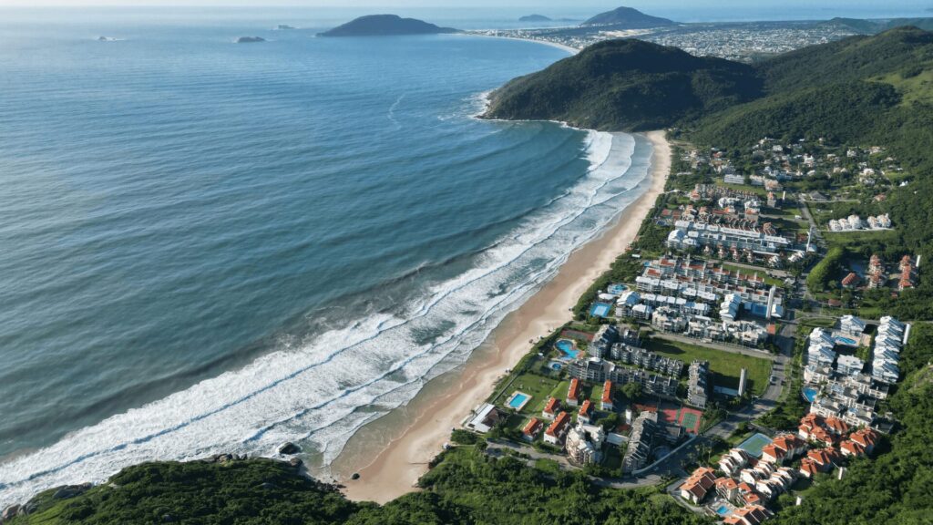 Vista aérea de uma bela praia com ondas ondulantes, cercada por colinas verdejantes e áreas residenciais, sob um céu azul claro.