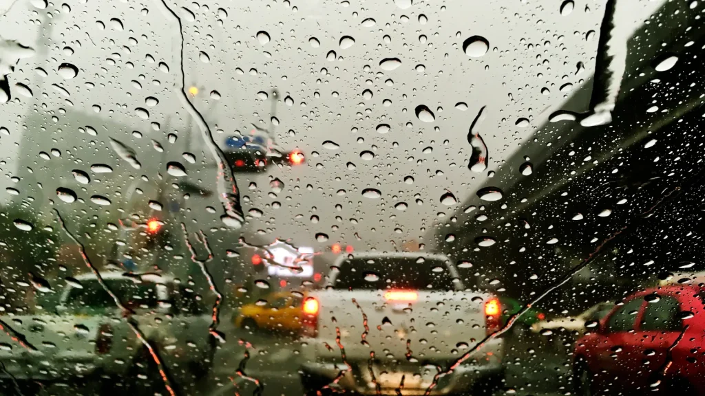 Gotas de chuva grudam no para-brisa de um carro, borrando a movimentada cena de uma rua da cidade com luzes vermelhas e amarelas dos veículos e céu nublado.