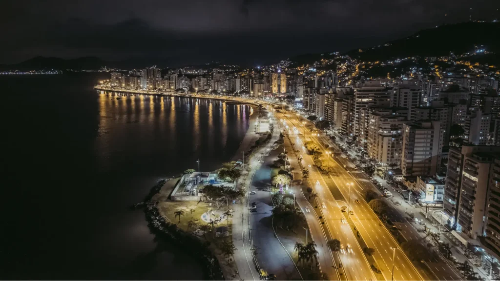 O horizonte de uma cidade à noite com edifícios iluminados ao longo de uma estrada à beira-mar, refletindo luzes nas águas calmas abaixo.