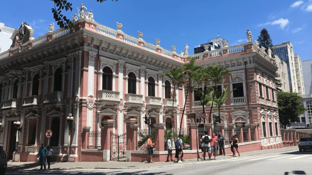 Edifício histórico rosa com arquitetura ornamentada, cercado por pedestres e palmeiras altas sob um céu azul claro.