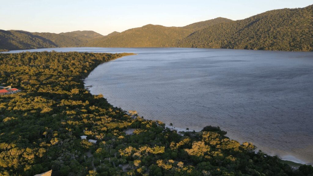 Vista aérea de um lago sereno cercado por florestas exuberantes e montanhas distantes sob um céu claro durante o pôr do sol.