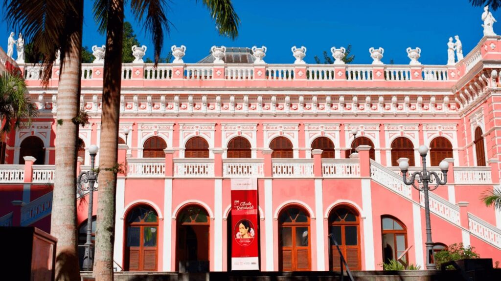 Uma foto de um grande edifício rosa-claro com detalhes brancos e telhado plano, adornado com balaustradas brancas e estátuas no topo. O edifício possui várias janelas de madeira escura com arcos no andar superior e algumas no andar inferior, além de escadas externas brancas em ambos os lados. Uma palmeira alta com tronco claro e folhas verdes pode ser vista no lado esquerdo da imagem, em primeiro plano, e uma bandeira vermelha está pendurada em uma das janelas do andar inferior.